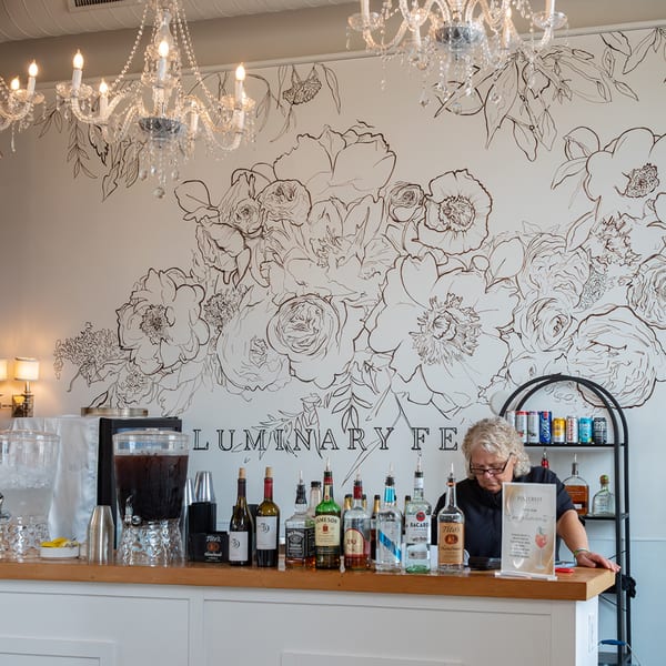 A Notch 8 bartender stands behind a bar with liquor bottles, drink dispensers, and chandeliers overhead.