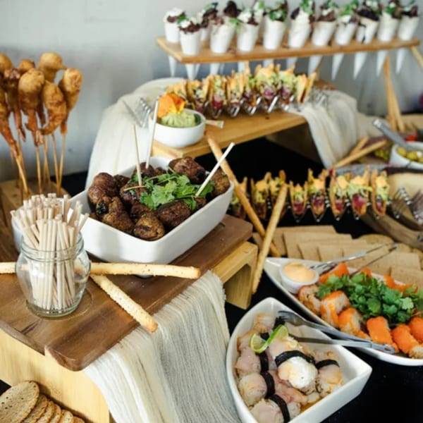 A buffet spread with various appetizers, including tacos, sushi, meatballs, and fried shrimp skewers.